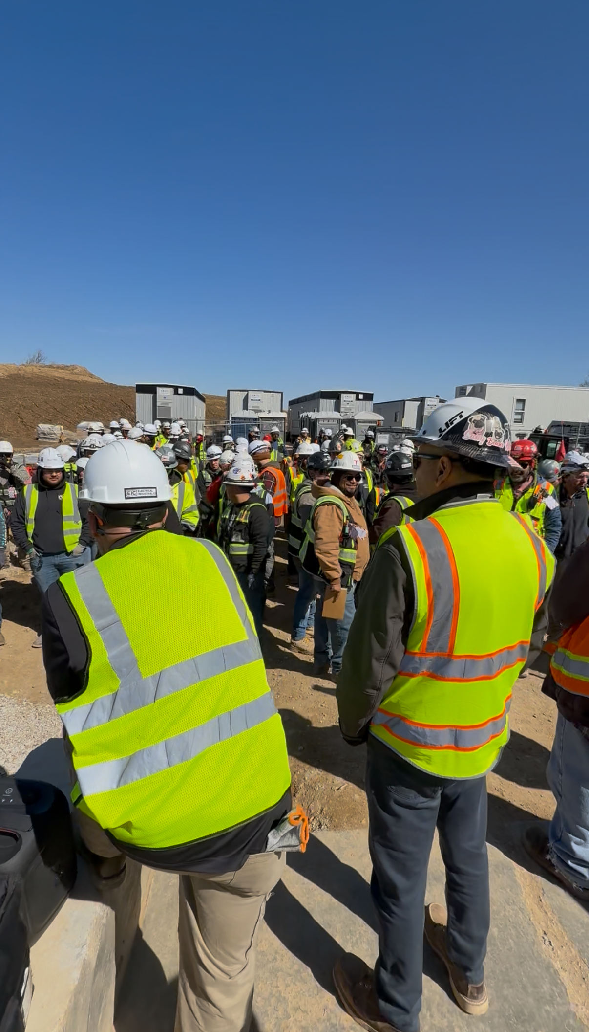 Construction field personnel gathered outdoors in hard hats and high-visibility vests.