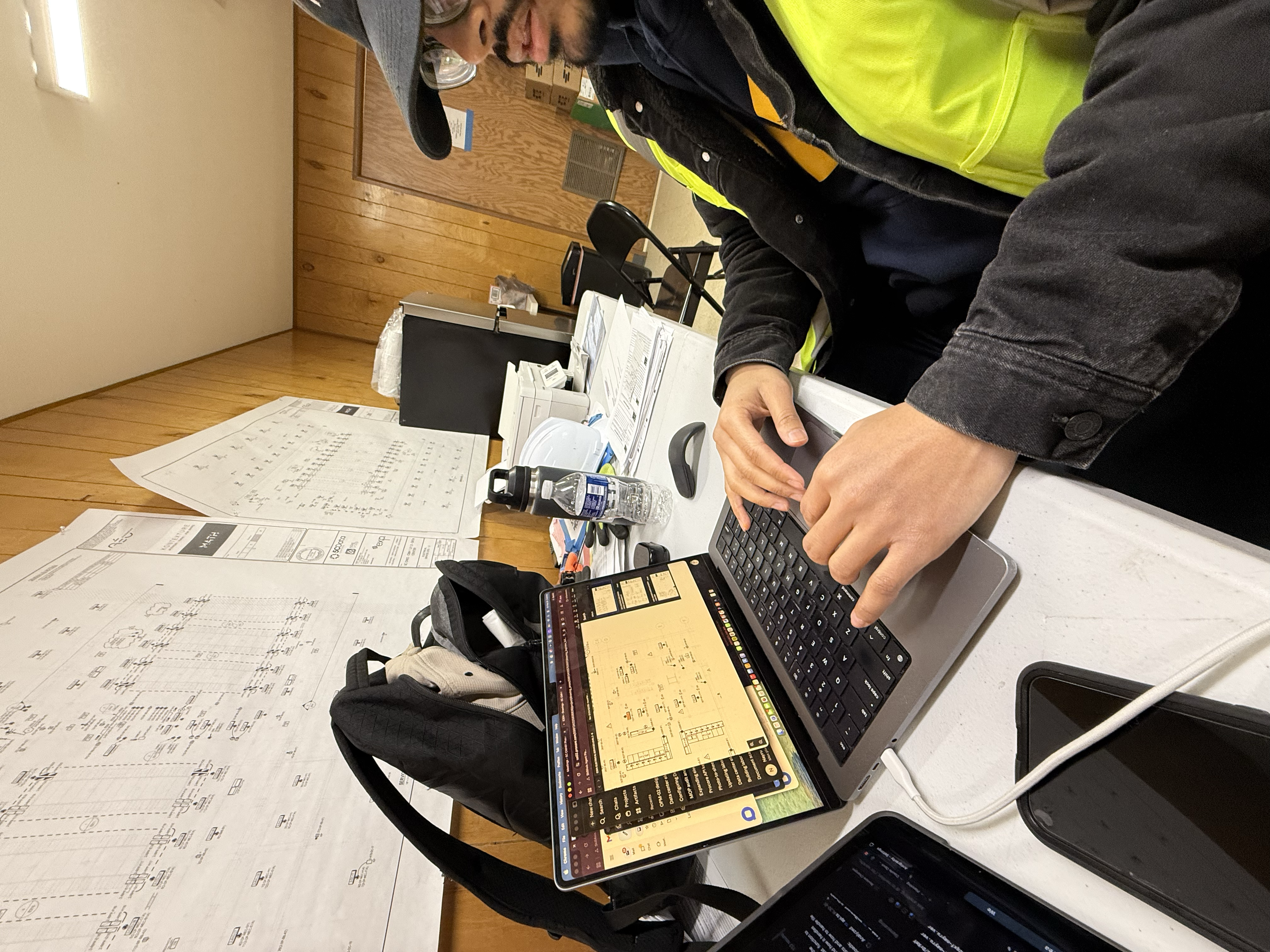Worker using a laptop at an onsite desk with electrical drawings on the wall.